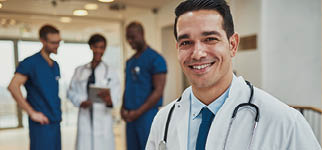 Successful handsome young Hispanic doctor with a beaming smile standing clutching a blue folder under his arm with his medical team in the background