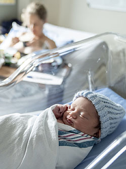 Un nouveau-né avec un bonnet bleu dans son berceau et sa maman dans le lit la maternité en second plan - MACSF