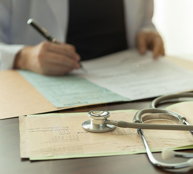 plan sur les mains d'un médecin qui remplit un dossier médical avec un stéthoscope posé sur le bureau - MACSF