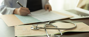 plan sur les mains d'un médecin qui remplit un dossier médical avec un stéthoscope posé sur le bureau - MACSF