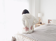 Une femme assise au bord de son lit se tenant le bas du dos - MACSF