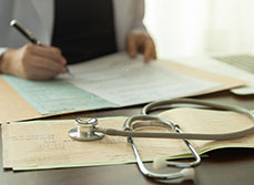 plan sur les mains d'un médecin qui remplit un dossier médical avec un stéthoscope posé sur le bureau - MACSF