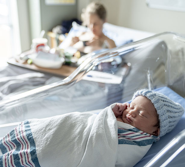 Un nouveau-né avec un bonnet bleu dans son berceau et sa maman dans le lit la maternité en second plan - MACSF