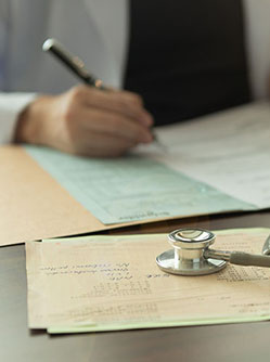 plan sur les mains d'un médecin qui remplit un dossier médical avec un stéthoscope posé sur le bureau - MACSF