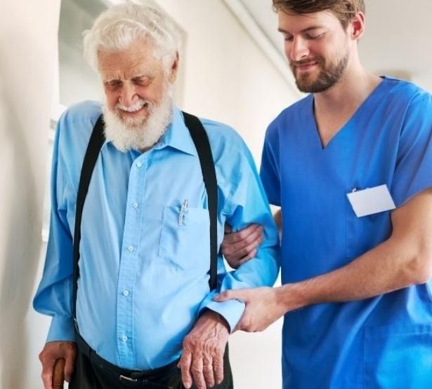 Un aide-soignant tient le bras d'un homme âgé pour l'aider à marcher dans le couloir de l'hôpital - MACSF