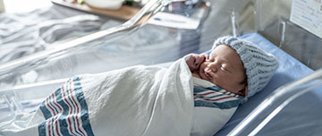 Un nouveau-né avec un bonnet bleu dans son berceau et sa maman dans le lit la maternité en second plan - MACSF
