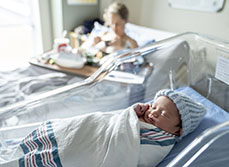 Un nouveau-né avec un bonnet bleu dans son berceau et sa maman dans le lit la maternité en second plan - MACSF