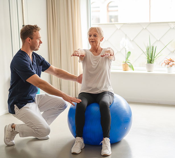 Un jeune kiné assiste une patiente âgée qui réalise des exercices d'équilibre à l'aide d'une Swiss Ball - MACSF