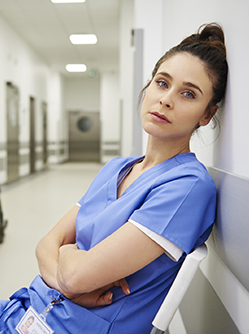 Une jeune professionnelle de santé à l'air triste assise et adossée au mur dans le couloir de l'hopital - MACSF