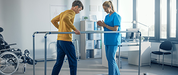 Un jeune homme marche en se tenant à des barres face à une kinésithérapeute - MACSF