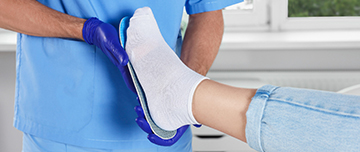 Un podologue en tenue bleue tient une semelle orthopédique contre le pied d’une personne assise - MACSF