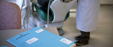 Un dossier patient posé sur un bureau au premier plan et un dentiste réalisant des soins à un patient sur le fauteuil dentaire en arrière plan - MACSF