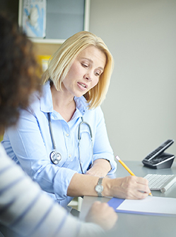 Une médecin assise à son bureau, en compagnie d'une patiente, rédige un document - MACSF