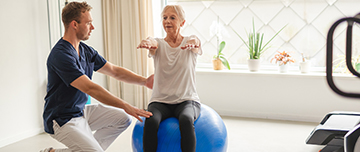 Un jeune kiné assiste une patiente âgée qui réalise des exercices d'équilibre à l'aide d'une Swiss Ball - MACSF