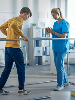 Un jeune homme marche en se tenant à des barres face à une kinésithérapeute - MACSF