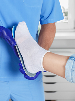 Un podologue en tenue bleue tient une semelle orthopédique contre le pied d’une personne assise - MACSF
