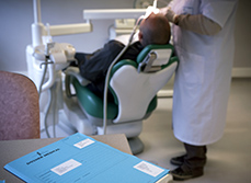 Un dossier patient posé sur un bureau au premier plan et un dentiste réalisant des soins à un patient sur le fauteuil dentaire en arrière plan - MACSF