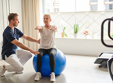 Un jeune kiné assiste une patiente âgée qui réalise des exercices d'équilibre à l'aide d'une Swiss Ball - MACSF