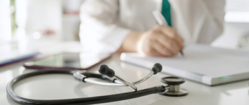 Un stéthoscope posé sur un bureau avec une professionnelle de santé qui prend des notes en arrière-plan - MACSF