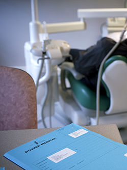 Un dossier patient posé sur un bureau au premier plan et un dentiste réalisant des soins à un patient sur le fauteuil dentaire en arrière plan - MACSF