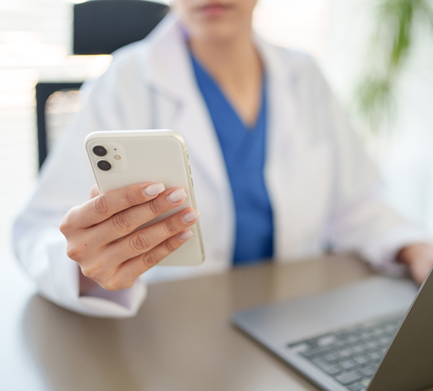 Gros plan sur la main d'une femme en blouse blanche tenant un téléphone portable et assise à son bureau devant un ordinateur - MACSF