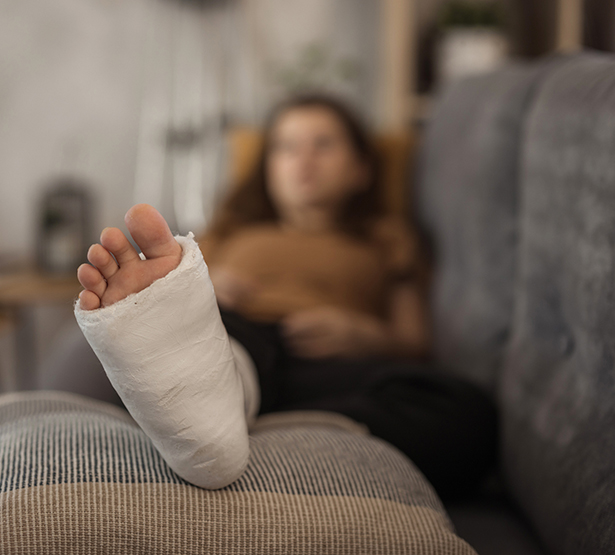 Gros plan sur le pied d'une femme en arrière plan à la jambe plâtrée allongée dans son canapé - MACSF
