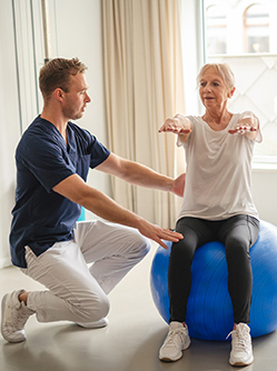Un jeune kiné assiste une patiente âgée qui réalise des exercices d'équilibre à l'aide d'une Swiss Ball - MACSF