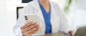 Gros plan sur la main d'une femme en blouse blanche tenant un téléphone portable et assise à son bureau devant un ordinateur - MACSF
