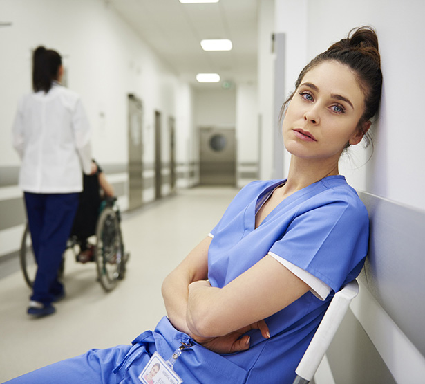 Une jeune professionnelle de santé à l'air triste assise et adossée au mur dans le couloir de l'hopital - MACSF