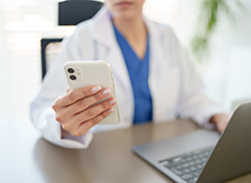 Gros plan sur la main d'une femme en blouse blanche tenant un téléphone portable et assise à son bureau devant un ordinateur - MACSF