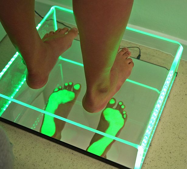 Gros plan sur des pieds nus posés sur une plateforme en verre éclairée en vert, avec empreintes visibles dessous. - MACSF