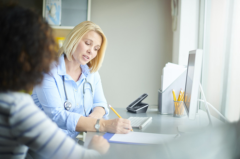Une médecin assise à son bureau, en compagnie d'une patiente, rédige un document - MACSF