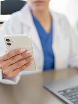 Gros plan sur la main d'une femme en blouse blanche tenant un téléphone portable et assise à son bureau devant un ordinateur - MACSF