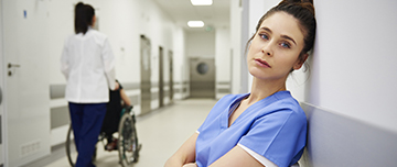 Une jeune professionnelle de santé à l'air triste assise et adossée au mur dans le couloir de l'hopital - MACSF