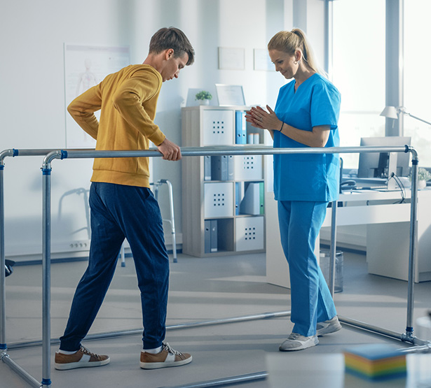 Un jeune homme marche en se tenant à des barres face à une kinésithérapeute - MACSF