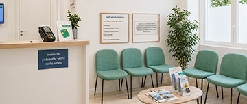 Une salle d’attente moderne avec des chaises vertes, une petite table, un comptoir d’accueil et des panneaux affichant les tarifs de consultation - MACSF