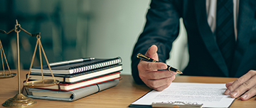 Gros plan sur une main d'homme s'apprêtant à écrire, avec à droite une pile de carnets et une balance de la justice - MACSF