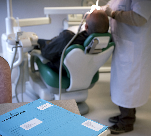 Un dossier patient posé sur un bureau au premier plan et un dentiste réalisant des soins à un patient sur le fauteuil dentaire en arrière plan - MACSF