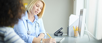 Une médecin assise à son bureau, en compagnie d'une patiente, rédige un document - MACSF